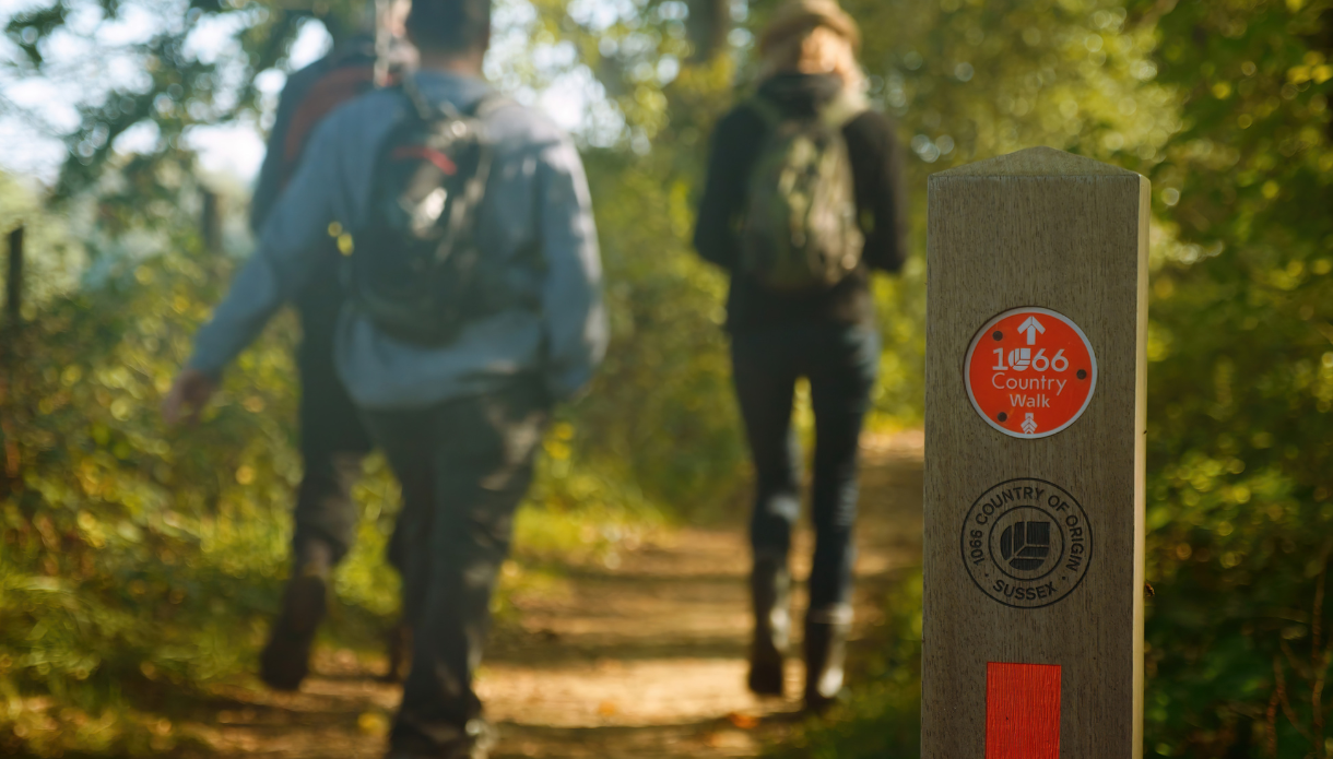 Two people walk along a wooded trail, with a 1066 Country Walk way marker in the foreground.